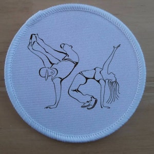 May include: White round patch with black line drawings of two people breakdancing. The man is doing a headspin and the woman is doing a handstand.