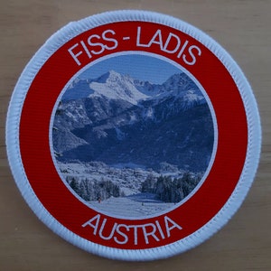 Könnte beinhalten: Ein weißer und roter runder Aufnäher mit dem Text "FISS-LADIS AUSTRIA" und einem Foto einer schneebedeckten Bergkette.