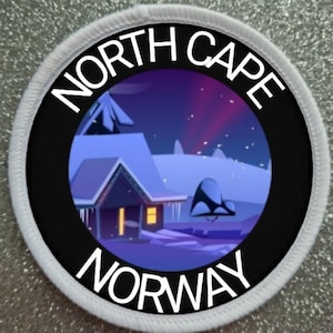 Puede incluir: Un parche circular blanco con texto negro que dice "NORTH CAPE NORWAY". El centro del parche presenta un círculo negro con una ilustración colorida de un paisaje nevado con una casa y la aurora boreal en el cielo.