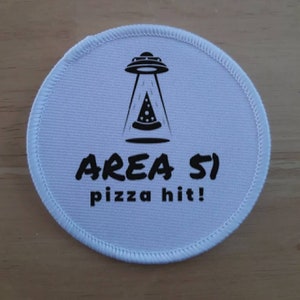 May include: White round patch with black text and a graphic of a UFO delivering a pizza. The text reads "AREA 51 pizza hit!"