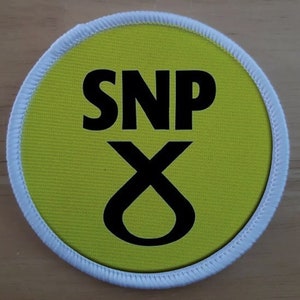 Może przedstawiać: Okrągły, żółty guzik z białym obszyciem wokół krawędzi. Guzik ma czarny napis "SNP" i czarny kształt "X".