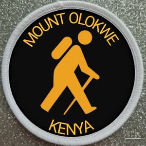 May include: A black and white round patch with a white border. The patch features a yellow silhouette of a hiker with a backpack and walking stick. The text "MOUNT OLOKWE KENYA" is written in yellow around the hiker.