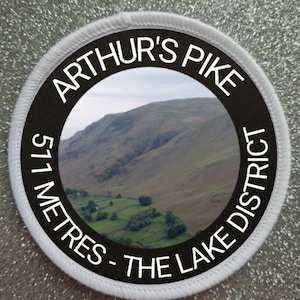 May include: A white and black round patch with a photo of a mountain range and the text "Arthur's Pike 571 Metres - The Lake District".
