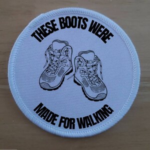 May include: White round patch with black text and a graphic of two hiking boots. The text reads "These boots were made for walking."