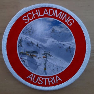 May include: White round patch with a red border. The patch has a picture of a snowy mountain scene with a ski lift in the center. The text "SCHLADMING AUSTRIA" is printed around the border.