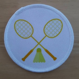 May include: A white round patch with a yellow badminton racket and shuttlecock design.