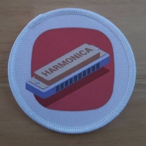 May include: Round patch with a white border and a red center. The patch features a blue and tan harmonica with the word "HARMONICA" printed on it. The patch is likely an iron-on or sew-on.