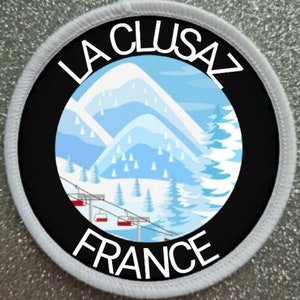 May include: A white and black round patch with a white border. The center of the patch is black with a white graphic of a mountain range with a ski lift. The text "LA CLUSAZ FRANCE" is written in white around the graphic.