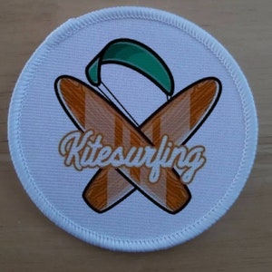 Peut inclure: Patch rond blanc avec un motif de kitesurf vert, blanc et orange. Le motif représente deux planches de surf croisées avec un cerf-volant au-dessus. Les mots "Kitesurfing" sont écrits en blanc, en cursive.