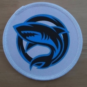 May include: A white round patch with a blue and black shark graphic. The shark is stylized and has a black outline with a blue fill. The shark is jumping out of the water and has its mouth open.