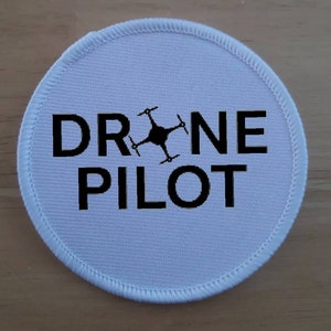 Könnte beinhalten: Runder weißer Aufnäher mit schwarzem Text "DRONE PILOT" und einer schwarzen Silhouette einer Drohne in der Mitte.