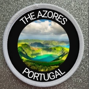 May include: A white circular patch with black text and a black circle in the center. The text reads "THE AZORES PORTUGAL". The black circle contains a photo of a green volcanic crater lake.