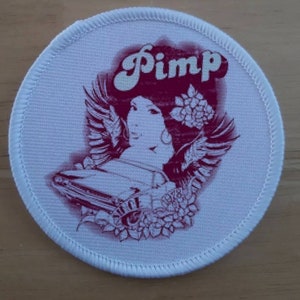 May include: White round patch with a pink and white illustration of a woman with wings, a car, and flowers. The text "Pimp" is written in a stylized font above the woman's head.