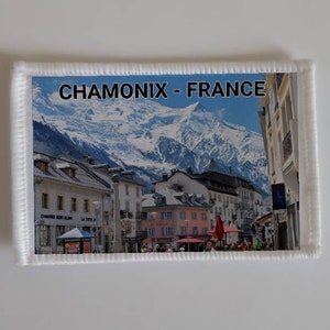 Könnte beinhalten: Ein weißer Patch mit einer gestickten Umrandung, der ein farbenfrohes Bild einer Stadt in den französischen Alpen zeigt. Der Text "CHAMONIX - FRANCE" ist oben auf dem Patch in Schwarz gedruckt.