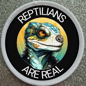 May include: A round white patch with black text that reads "Reptilians Are Real". The center of the patch features a cartoon image of a green and yellow lizard with orange eyes.