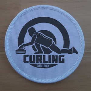 Può includere: Un cerotto rotondo bianco con una silhouette nera di un curler che fa scorrere una pietra verso un bersaglio. Il testo "CURLING CHALLENGE" è stampato sotto l'immagine.