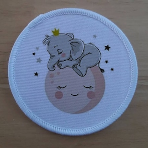 以下が含まれることがあります： 白い布地の円形のワッペンで、金色の冠をかぶった灰色の象が、笑顔のピンク色の月に寝ています。背景は白で、灰色の星があります。
