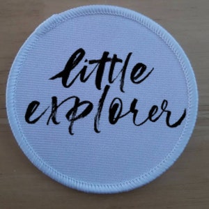 Op de afbeelding: Een ronde, lichtblauwe patch met de woorden "little explorer" in zwart script. De patch heeft een witte, gestikte rand. Geschikt voor kleding of accessoires.