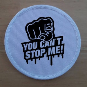 May include: White round patch with black text and a black fist pointing at the viewer. The text reads "You Can't Stop Me!"