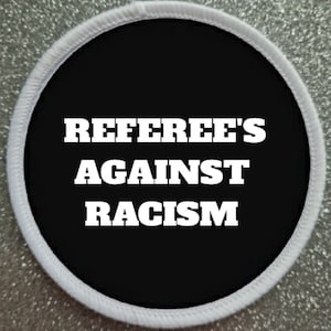 Könnte beinhalten: Ein runder Aufnäher mit weißem Rand und dem Text "REFEREE'S AGAINST RACISM" in fetten weißen Buchstaben auf schwarzem Hintergrund. Der Aufnäher ist wahrscheinlich für Kleidung oder Accessoires.
