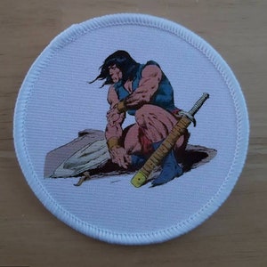 Peut inclure: Un écusson rond blanc avec une image colorée d'une figure musclée tenant une épée. La figure est à genoux et a de longs cheveux noirs. L'arrière-plan est de couleur brun clair.