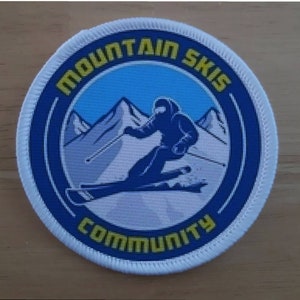 Peut inclure: Un écusson rond blanc avec une bordure bleue et du texte jaune. Le texte indique "Mountain Ski Community". À l'intérieur du cercle, une silhouette bleue d'un skieur sur une montagne blanche avec un fond de ciel bleu.
