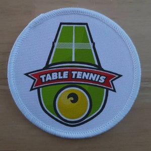 Könnte beinhalten: Weißer runder Patch mit einem grünen Tischtennis-Schläger und einem gelben Ball. Der Schläger hat ein weißes Netz und den Text "TABLE TENNIS" in roten Buchstaben auf einem roten Band.