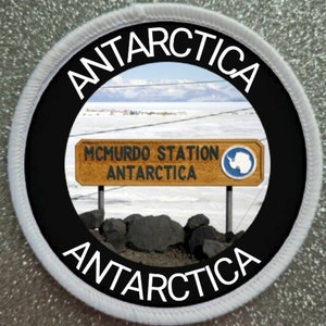 May include: Circular patch with the word "ANTARCTICA" in white lettering. The centre shows a sign for McMurdo Station, Antarctica, with a snowy landscape and mountains in the background.