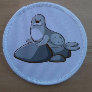 Può includere: Una toppa di tessuto bianco con una foca grigia seduta su uno scoglio grigio. La foca ha gli occhi neri e il naso nero.