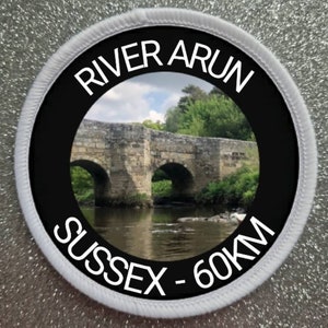 May include: A white round patch with black text and a photo of a stone bridge over a river. The text reads "River Arun Sussex - 60km".