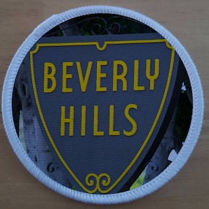 May include: Round patch featuring the text "BEVERLY HILLS" in yellow letters on a gray shield-shaped sign. The sign has a yellow border and decorative swirls. The patch is framed by a white stitched border.