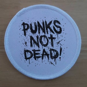 Peut inclure: Écusson rond en tissu blanc avec du texte noir qui dit "Punks Not Dead!" dans une police de style graffiti qui coule.