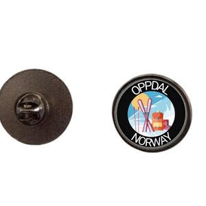 May include: A round metal pin featuring a graphic of skis, a backpack, and snowy mountains with the text OPPDAL NORWAY. The pin has a black border and a dark metallic back with a butterfly clutch fastener for secure attachment.