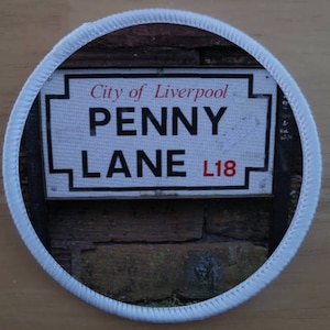 Può includere: Toppa rotonda con un cartello stradale di Penny Lane a Liverpool. Il cartello recita "City of Liverpool" sopra "PENNY LANE L18" in lettere nere e rosse. La toppa ha un bordo bianco.