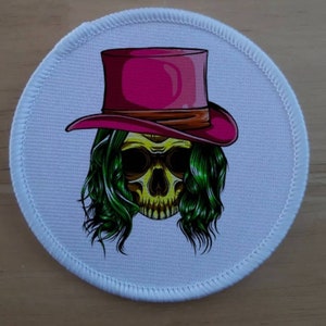May include: White round fabric patch with a skull wearing a pink top hat with a brown band. The skull has green hair and yellow eyes.