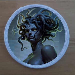 May include: A round patch with a white border featuring a detailed illustration of Medusa, a mythical Greek figure with snakes for hair, a fierce expression, and piercing yellow eyes.