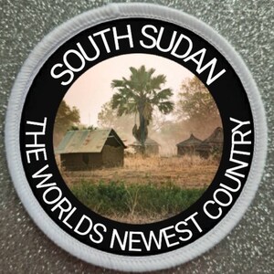 May include: A black and white circular patch with the text "South Sudan The Worlds Newest Country" around the edge. The center of the patch features a photograph of a village in South Sudan with a palm tree in the center.