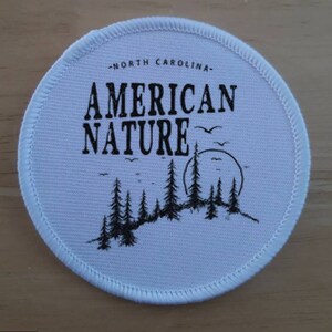 Könnte beinhalten: Ein weißer runder Patch mit schwarzen Nähten am Rand. Der Patch hat schwarzen Text, der "North Carolina - American Nature" lautet, und eine schwarze Silhouette eines Waldes mit einer Sonne und Vögeln.