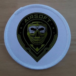 以下が含まれることがあります： 黒い円の中に「AIRSOFT」という文字が入った丸い白いパッチ。パッチには、黄色と黒のマスクをつけたスカルが描かれています。パッチには白い縁取りがあります。
