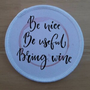 Pode incluir: Patch redondo de tecido branco com uma mancha de vinho rosa no centro. O texto no patch diz "Be nice Be useful Bring wine".