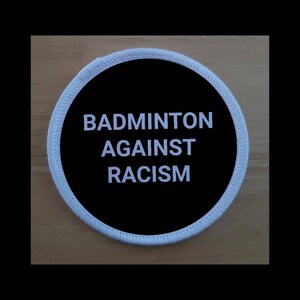 Può includere: Un cerotto rotondo nero e bianco con il testo "Badminton Against Racism" in bianco.