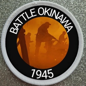 May include: A black and white circular patch with the text "BATTLE OKINAWA 1945" in white. The center of the patch features a silhouette of soldiers in a foggy orange landscape.