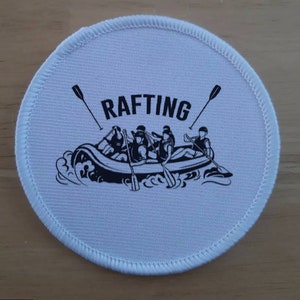 Puede incluir: Un parche redondo blanco con costuras negras alrededor del borde. El parche presenta una ilustración en blanco y negro de una balsa con cinco personas remando por un río. El texto "RAFTING" está impreso en negro sobre la balsa.