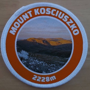 Könnte beinhalten: Ein runder weißer und orangefarbener Aufnäher mit dem Text "MOUNT KOSCIUSZKO" in einem weißen Kreis um ein Foto einer Bergkette. Der Text "2228m" ist unter dem Foto gedruckt.