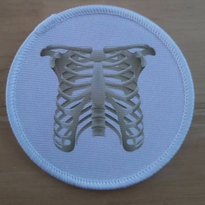 May include: White round fabric patch with a gray image of a human rib cage.