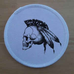 Op de afbeelding: Een ronde witte patch met een zwart-witte illustratie van een schedel met een mohawk en veren.