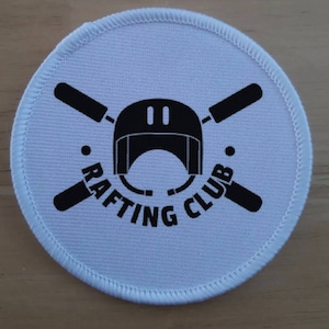 Puede incluir: Parche redondo blanco con texto negro y un gráfico de un casco con remos cruzados. El texto dice "Rafting Club".