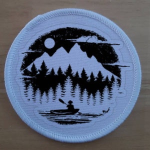 Könnte beinhalten: Runder weißer Patch mit einer schwarzen Silhouette einer Bergkette, Bäumen und einer Person in einem Kanu auf einem See. Der Mond ist am Himmel sichtbar.