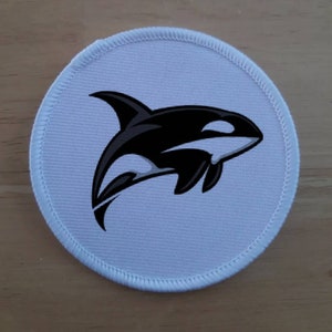 Puede incluir: Parche redondo blanco con un gráfico de orca en blanco y negro. La orca está nadando con la boca abierta y una mancha blanca en el ojo.