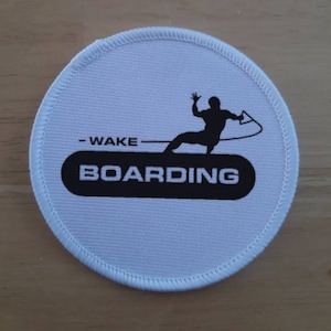 Puede incluir: Parche redondo blanco con costuras negras alrededor del borde. El parche presenta una silueta negra de una persona haciendo wakeboard con el texto "WAKE BOARDING" en negro.
